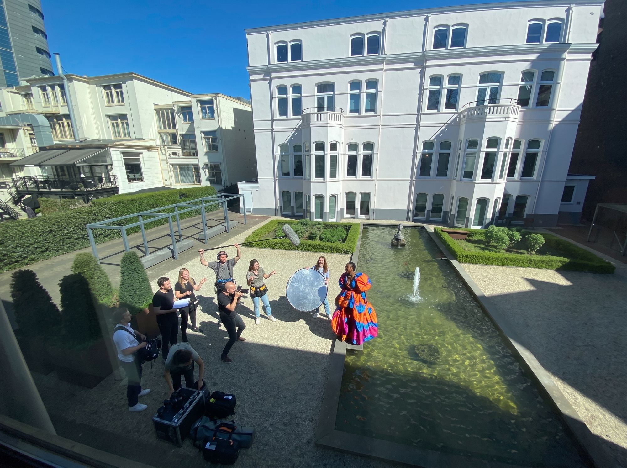 Fotoshoot in de tuin, gezien vanuit de verdieping: een crew met camera en lichtkoffers en een model in een kleurige jurk naast de waterpartij