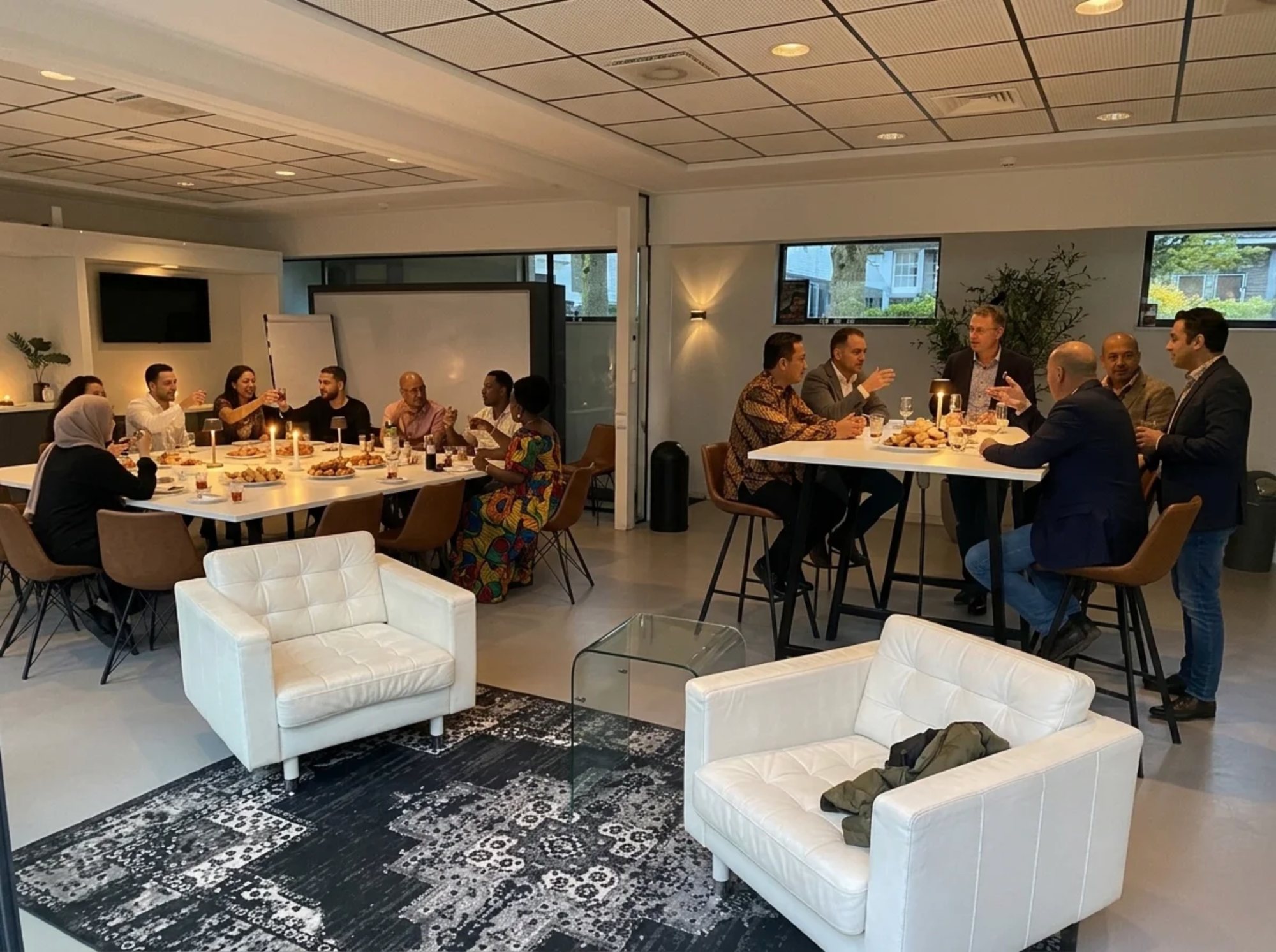 Een gemengd gezelschap proost met thee en wijn aan de lange tafel in de zaal bij kaarslicht, met gebak en schalen op tafel en aan de statafel een gesprek in het avondlicht
